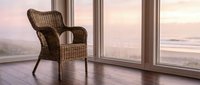 Woven rattan armchair positioned by expansive floor-to-ceiling windows overlooking serene coastal landscape at sunset, creating peaceful interior design ambiance.