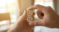 Hands holding wooden tokens with happy and sad face emoticons representing emotional choice and mental health awareness in warm indoor setting.