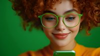 Young woman with curly red hair and green glasses smiling while looking at mobile phone screen against vibrant green background. Warm expression and modern style.