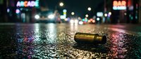 Empty bullet casing lies on wet urban street at night with neon arcade and bar signs glowing in blurred background, suitable for crime scene, noir, and gritty storytelling projects.
