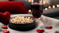 Cozy evening scene with popcorn bowl and red wine glass surrounded by heart decorations and warm bokeh lights for romantic date night at home.