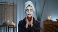 Young woman with white towel on head applying facial mask in spa bathroom with candles for skincare routine and relaxation at home wellness treatment.