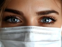 Close up view of expressive blue eyes with long eyelashes and well-groomed eyebrows above protective medical face mask, representing healthcare and safety during pandemic.