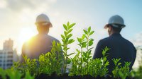 Two construction workers in hard hats silhouetted against sunset sky with green plants in foreground, representing sustainable development and eco-friendly building concept.