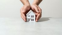 Male hands protecting small white house model on clean surface symbolizing home insurance security and real estate protection concept.