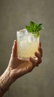 Hand holding refreshing cocktail with mint garnish and ice cubes in crystal glass against neutral background for beverage and hospitality concepts.