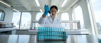 Asian female scientist in white lab coat examining blue test tubes in modern laboratory with city skyline view through windows for research analysis.