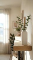 Minimalist home interior with white ceramic vase holding eucalyptus branches and wooden sticks on wooden shelf, bright natural light from window, neutral beige walls, scandinavian style decor.