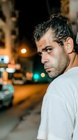 Male model with dark hair and Mediterranean ethnicity wearing white t-shirt, looking over shoulder at night urban street with blurred city lights and traffic in background.