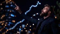 Caucasian bearded businessman in dark coat pointing up at night city lights with bokeh effect background, profile view showing confidence and vision.
