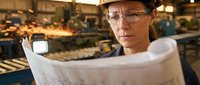 Middle-aged male engineer with glasses and hard hat reviewing construction blueprints in industrial factory setting with machinery and workers in background.