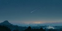 Meteor streaking across twilight sky over misty mountain landscape with city lights glowing on horizon creating serene evening atmosphere for nature photography.
