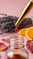 Golden dropper releasing amber serum drop into glass bottle, lavender and orange slices on pink background, natural skincare concept.