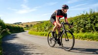 Professional cyclist in aerodynamic position racing on rural road through green countryside landscape during training session for competitive sports.