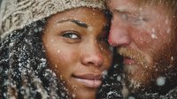 Young mixed-race woman and Caucasian man sharing intimate winter moment with snowflakes falling, cozy knit hat, romantic couple outdoors in cold weather.