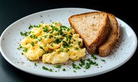 Fluffy scrambled eggs garnished with fresh chopped chives served alongside golden-brown toast on white plate against dark background. Classic breakfast presentation.