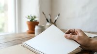 Close-up of hand holding blank spiral notebook on wooden desk with potted plant and pencil holder, suitable for stationery, office, education, and creative workspace concepts.