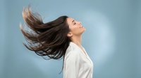 Young woman with flowing brown hair in motion expressing joy and freedom against soft blue background for wellness and lifestyle concepts.