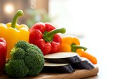 Fresh colorful vegetables including broccoli, bell peppers, eggplant and carrots arranged on wooden cutting board in bright kitchen for healthy cooking.