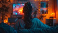 Young woman relaxing on couch watching TV in dark room with ambient orange mood lighting and bokeh effects, back view of messy bun hairstyle at night.