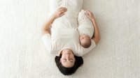 Happy pregnant Asian woman lying on carpet with newborn baby in white clothing bonding together in peaceful maternity moment at home.