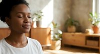 Young African American woman with eyes closed in peaceful meditation pose at home with natural lighting and plants in cozy living room interior.