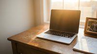 Modern laptop computer on wooden desk with warm sunlight streaming through window creating cozy home office workspace atmosphere for remote work and productivity.