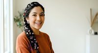 Young woman with long black braided hair and South Asian ethnicity wearing an orange-brown sweater, smiling at camera in bright modern interior with plants and white decor.