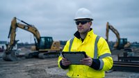 Construction site manager in high visibility safety gear using digital tablet with excavators in background for project management and planning.
