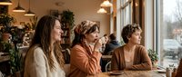 Three diverse women enjoying coffee together in cozy cafe with warm lighting and plants. Friends socializing, bonding over drinks in comfortable restaurant setting.