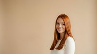 Young woman with long reddish-brown hair and fair complexion wearing white long-sleeve top, smiling at camera against neutral beige background for portrait and headshot use.