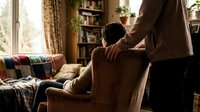 Caring partner providing comfort and support to loved one sitting in cozy living room chair, showing emotional connection and relationship intimacy.