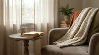 Cozy reading corner with knitted blanket draped over beige armchair, wooden side table with books and coffee cup, soft natural lighting from window.