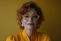 Mature Caucasian woman with curly red hair wearing bright orange glasses and matching yellow blouse smiles warmly against golden background. Professional portrait.