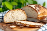 Fresh crusty bread loaf sliced on wooden cutting board with checkered napkin, showing soft white crumb texture and golden brown crust in natural light.