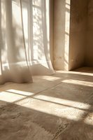 Soft morning sunlight casting window shadows through sheer white curtains onto textured beige floor, creating atmospheric play of light and shadow in minimal interior.