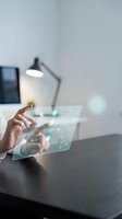 Hands of a person interacting with a transparent holographic tablet displaying digital data and interface elements at a modern desk workspace for technology and innovation concepts.