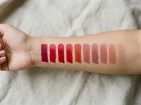 Woman's arm displaying colorful lipstick swatches from deep red to nude pink shades for makeup testing and beauty product comparison.