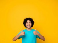 Young male with curly dark hair and medium brown skin tone wearing bright turquoise shirt giving double thumbs up gesture against vibrant yellow background, expressing approval and positivity for advertising campaigns.
