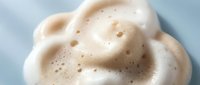 Creamy foam texture with bubbles on a light blue background, close-up macro shot of soft white lather for beauty and skincare concepts.