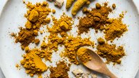 Turmeric powder scattered on white plate with wooden spoon, overhead view for cooking, wellness, and culinary content.