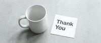 White ceramic mug beside a thank you note card on a gray concrete surface, conveying gratitude and appreciation in a minimal style.