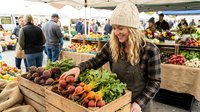 Young blonde woman in beige knit hat selecting fresh organic vegetables at bustling farmers market with wooden crates full of colorful produce.