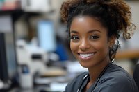 Young African American businesswoman with natural curly hair and bright smile in professional office setting, wearing black blazer against blurred workspace background.