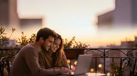 Young couple working together on laptop computer on romantic balcony terrace during golden hour sunset with city lights and plants in background.