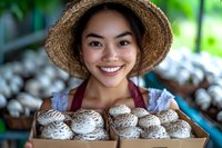 Young Asian woman farmer in straw hat and apron holding wooden box of fresh harvested mushrooms, smiling at camera against blurred greenhouse background.