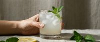 Caucasian hand holding a glass of icy lemonade with fresh mint garnish on a linen surface, with lemon slices nearby.