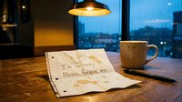 Handwritten apology note on wooden table with coffee stains, mug, and pen under warm lamp light, rainy window background, emotional concept.