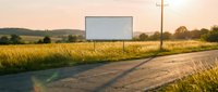 Blank white billboard beside a rural road at golden sunset, surrounded by green fields, ready for advertising or promotional mockup design.