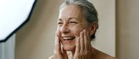 Mature woman with gray hair and fair complexion smiling while touching her face, expressing joy and skincare satisfaction for wellness and beauty campaigns.